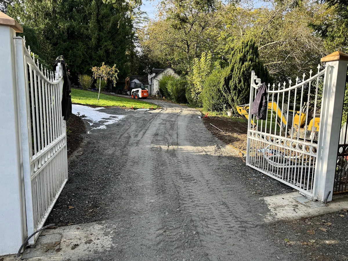 Allée décaissée entre deux battants de portail, mini-pelle au travail en arrière-plan