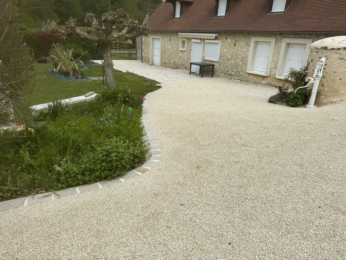 Grande cour en gravier clair devant une longère, délimitée par une bordure de pavés courbe autour d'un massif