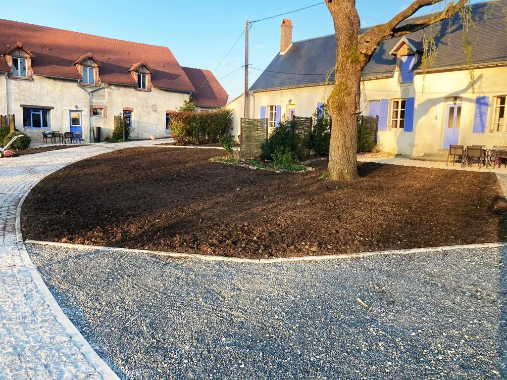 Cour en gravier stabilisé bordant deux longères, avec un massif de terre fraîchement préparée sous un grand arbre