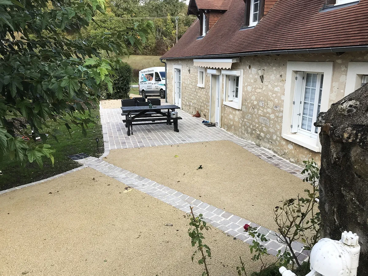 Vue d'ensemble d'une cour terminée, terrasse pavée et surfaces en gravier devant une longère