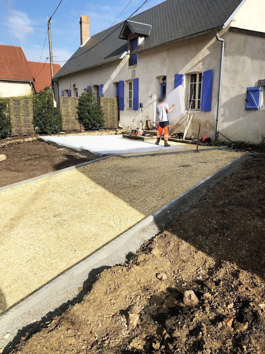 Compagnon déployant le géotextile sur une cour en préparation, bordures déjà en place
