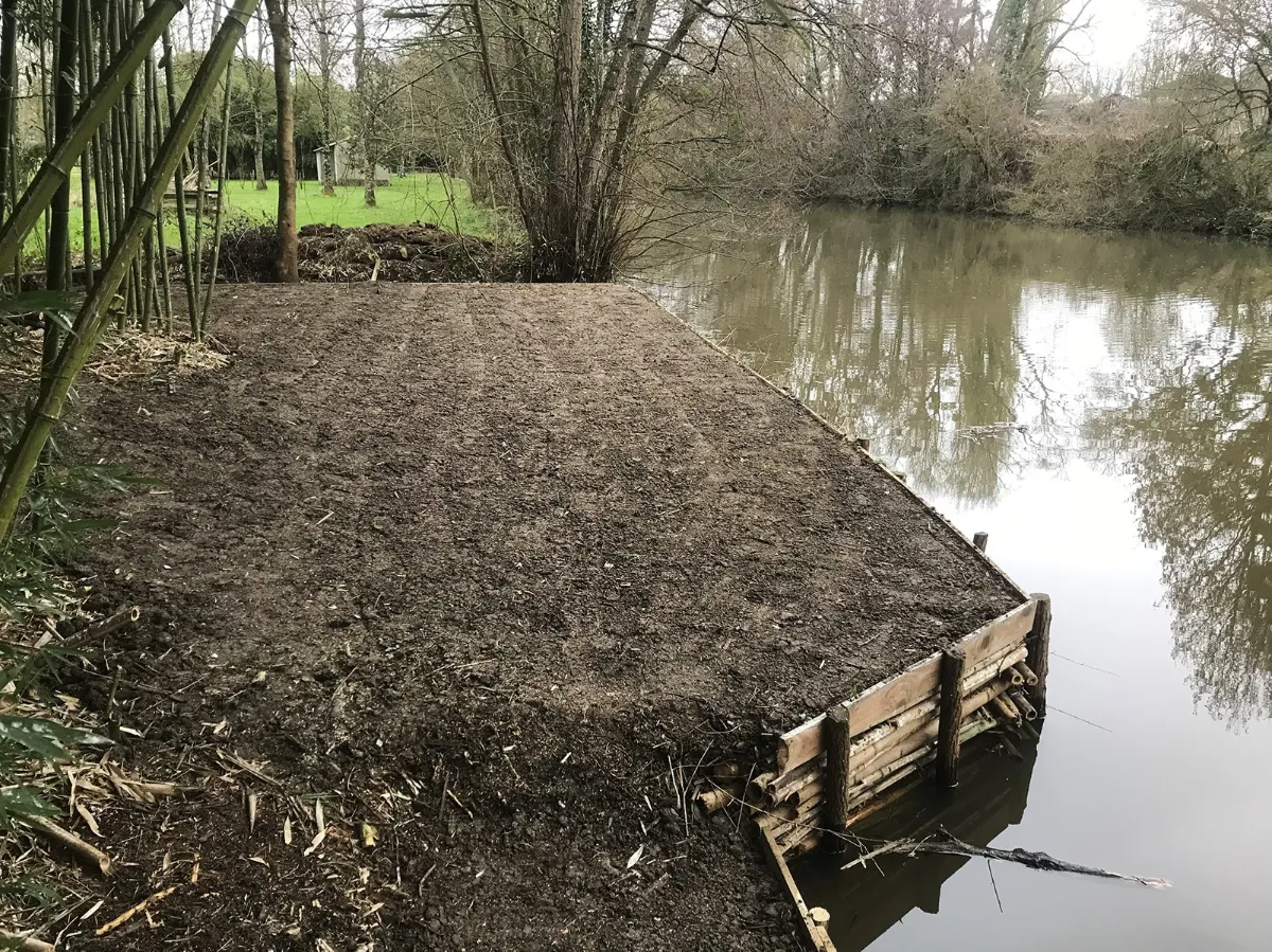 Plateforme de terre nivelée derrière un tunage de rondins, en bordure de rivière