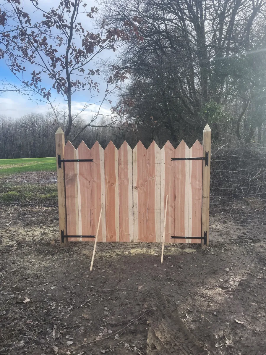 Portail de bois neuf à lames pointues, monté entre deux poteaux, en bordure de champ