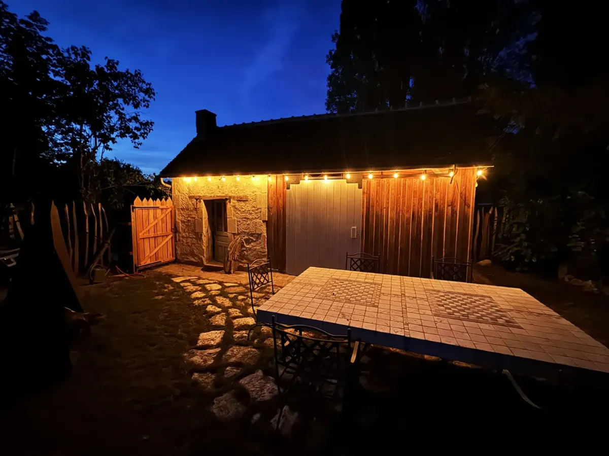 Appentis en bardage bois accolé à une longère de pierre, terrasse et guirlandes lumineuses au crépuscule