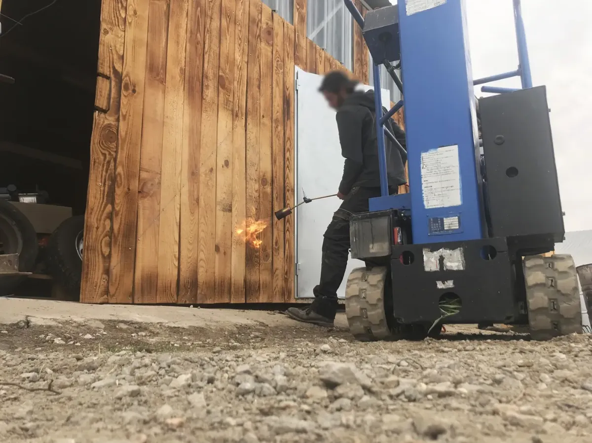 Flamme du chalumeau léchant les lames du bardage d'un hangar, compagnon à l'œuvre depuis une nacelle