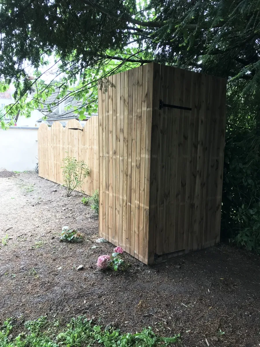 Petit cabanon en bardage bois avec porte pleine, devant une palissade à l'arase arrondie