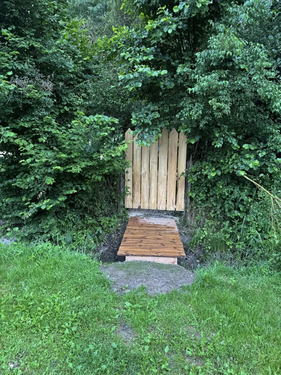 Petite passerelle en bois franchissant un fossé et menant à un portillon ouvert dans une haie