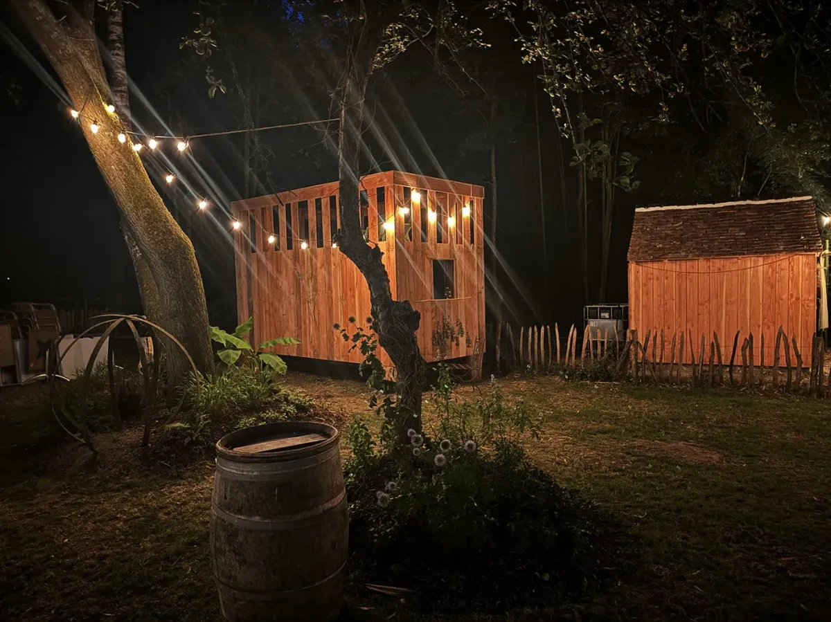 Pavillon de jardin en bois éclairé de guirlandes à la nuit tombée, abri de jardin voisin