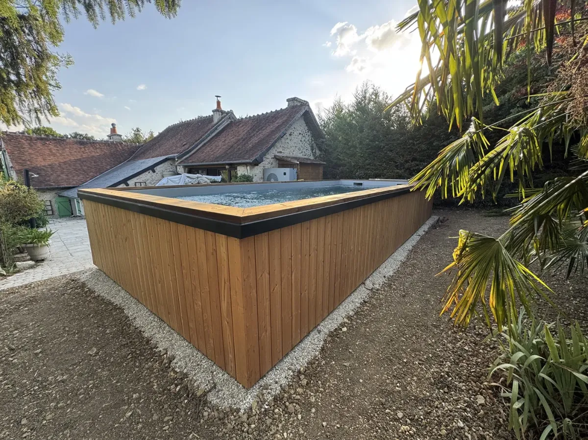 Piscine hors-sol en bois dans un jardin de campagne, toits d'une longère en arrière-plan