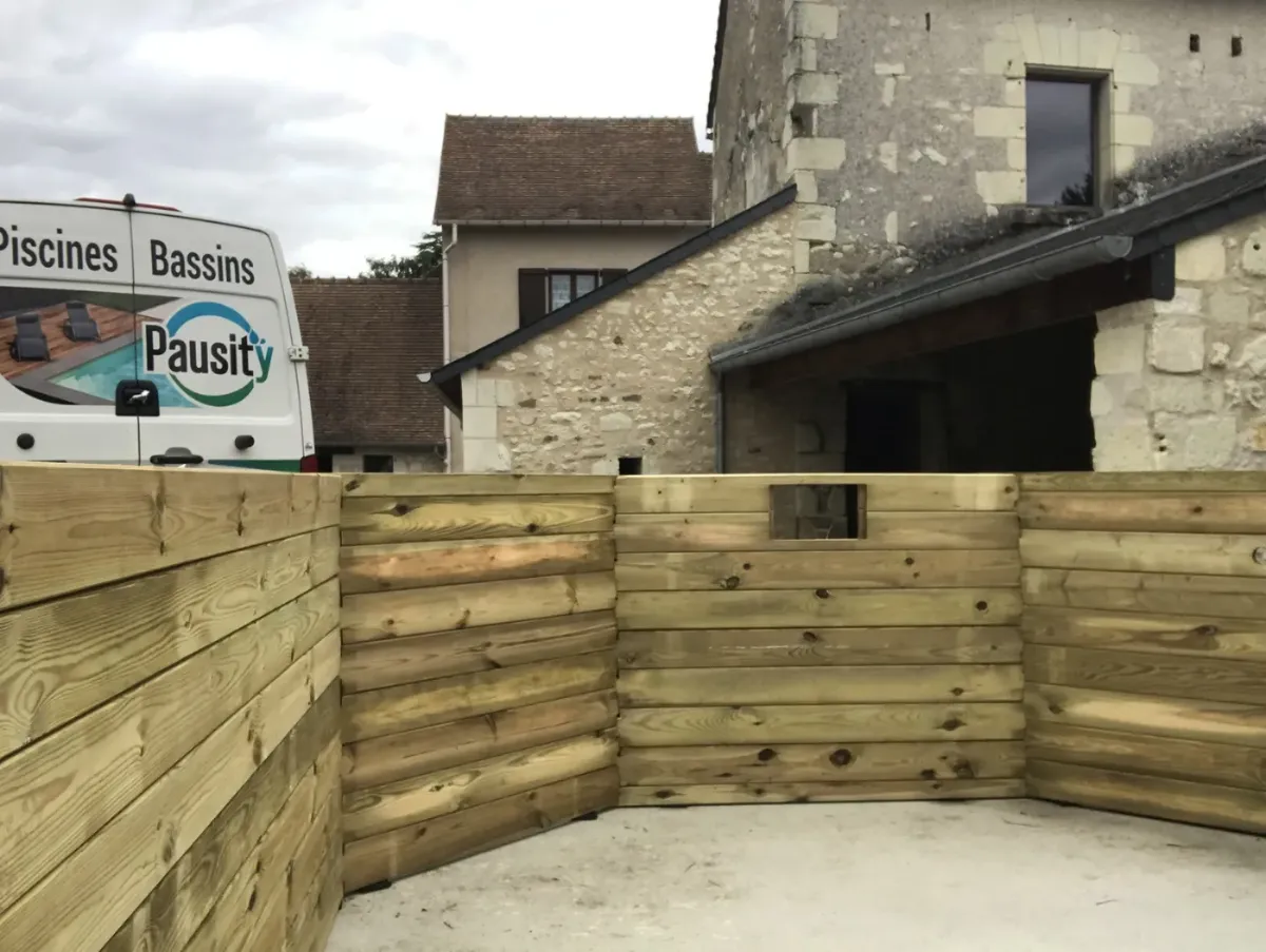 Piscine bois montée sur dalle béton, ouverture du skimmer visible dans les madriers, ferme en pierre au fond