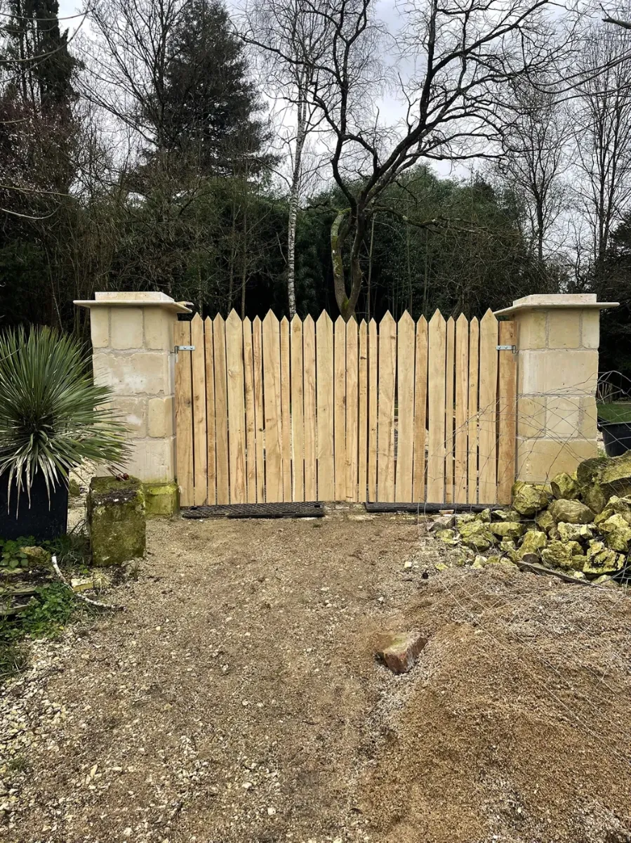 Portail de jardin en lames de bois pointues entre piliers de pierre, avant la pose du cadre contreventé