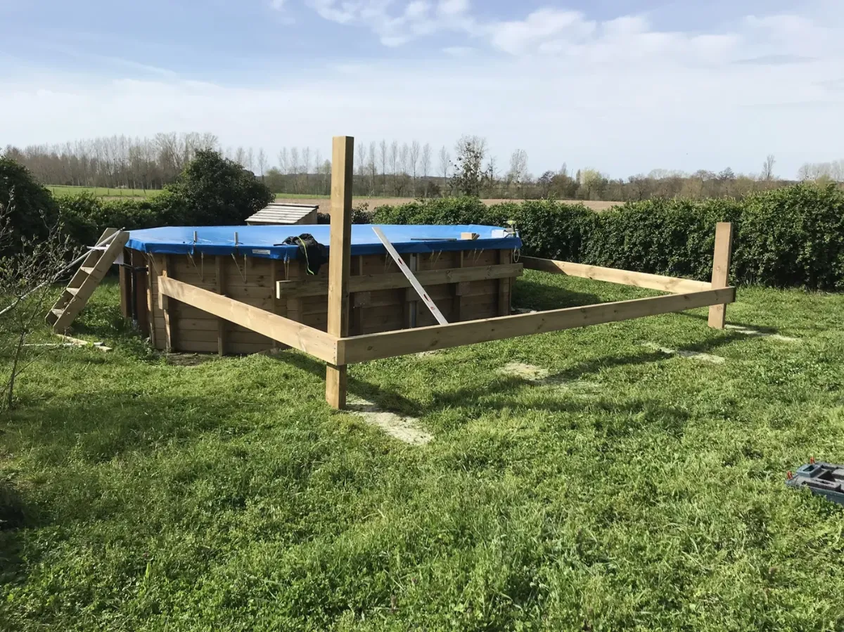 Poteaux et lisses d'implantation d'une terrasse de piscine plantés dans l'herbe, piscine bâchée derrière