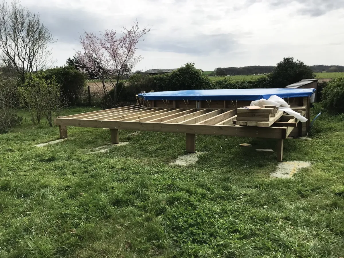 Structure de terrasse en solives de bois montée sur plots, contre une piscine hors-sol bâchée