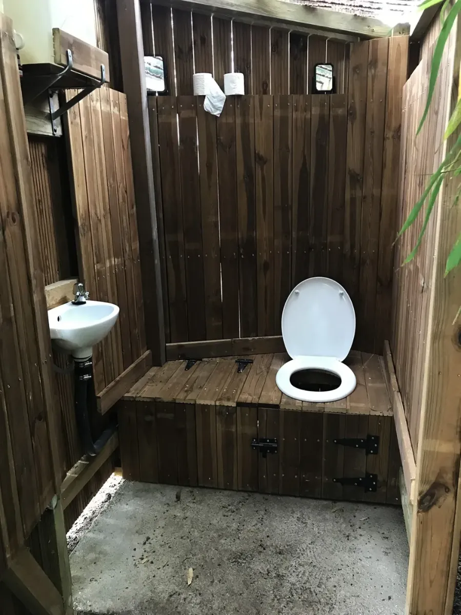 Toilettes sèches habillées de bois, avec abattant et petit lave-mains, éclairées par une fenêtre