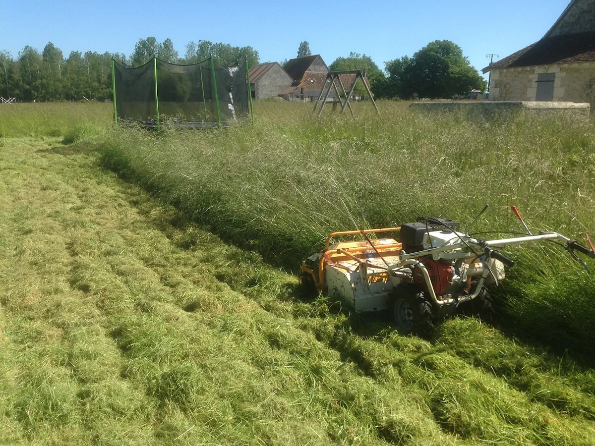 Débroussailleuse autotractée fauchant une prairie haute devant une longère, large andain d'herbe coupée sur la gauche