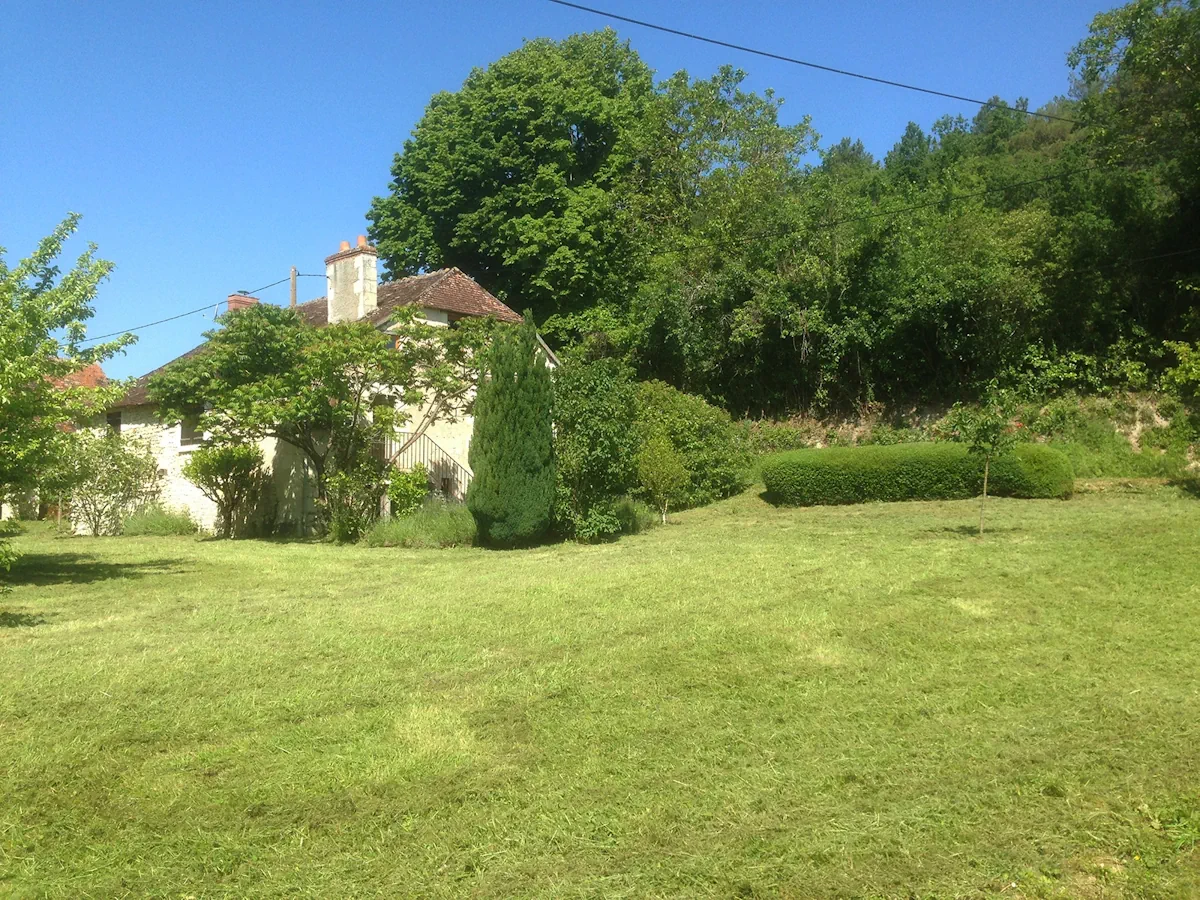 Jardin fauché autour d'une maison de pierre adossée à un coteau boisé