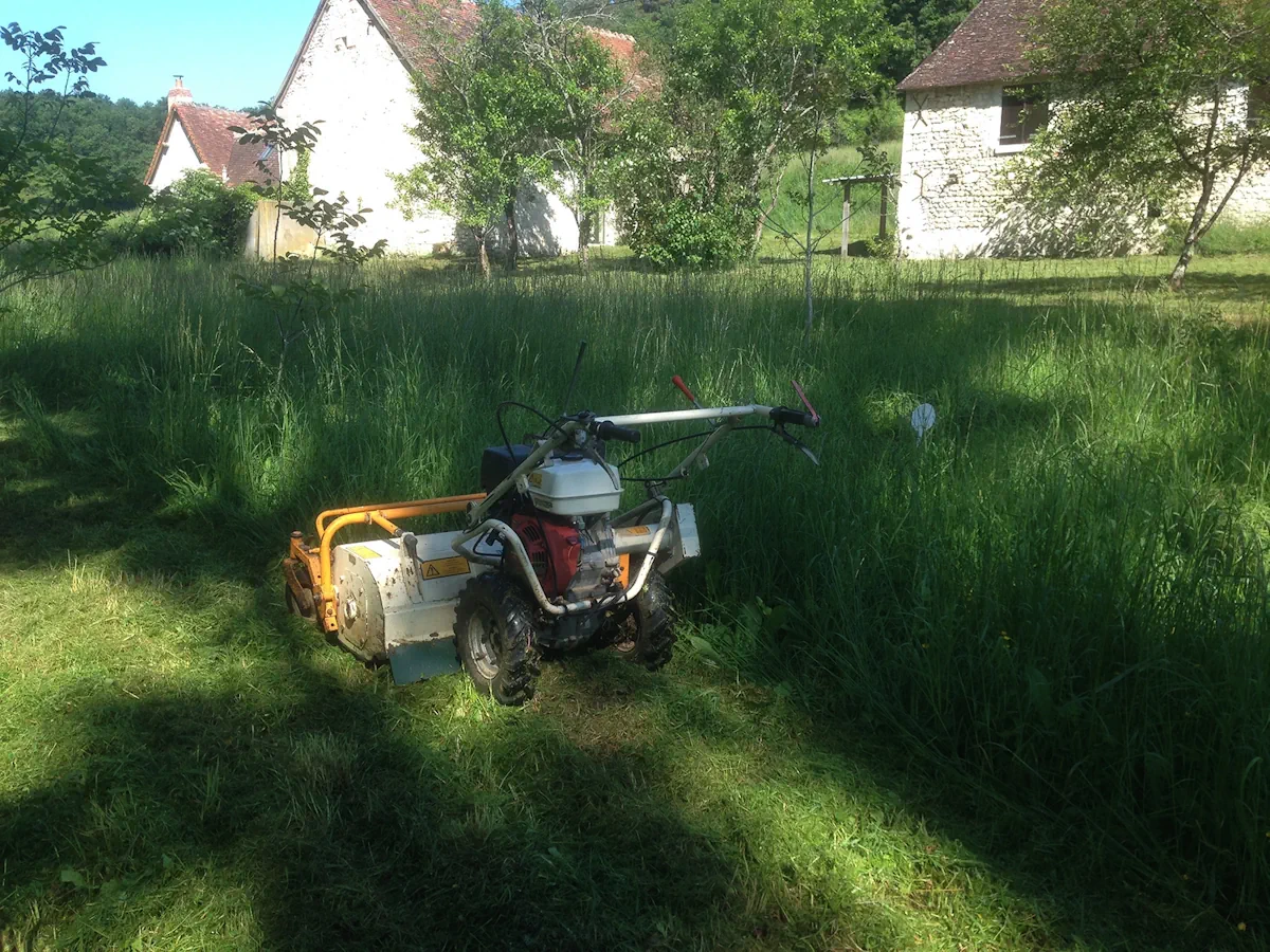 Débroussailleuse autotractée fauchant un verger enfriché devant des bâtiments de pierre