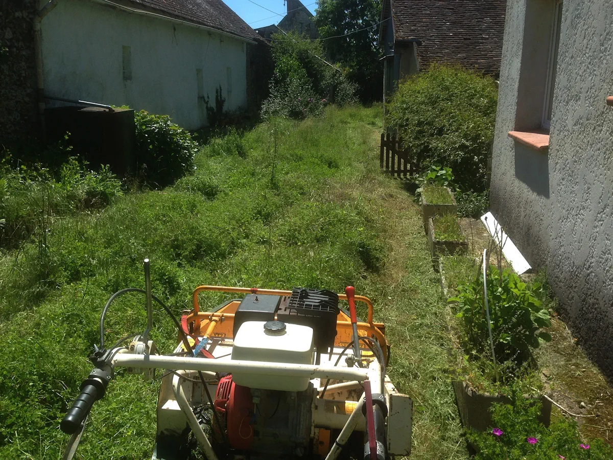 Passage entre deux bâtiments envahi de hautes herbes, débroussailleuse engagée à l'entrée