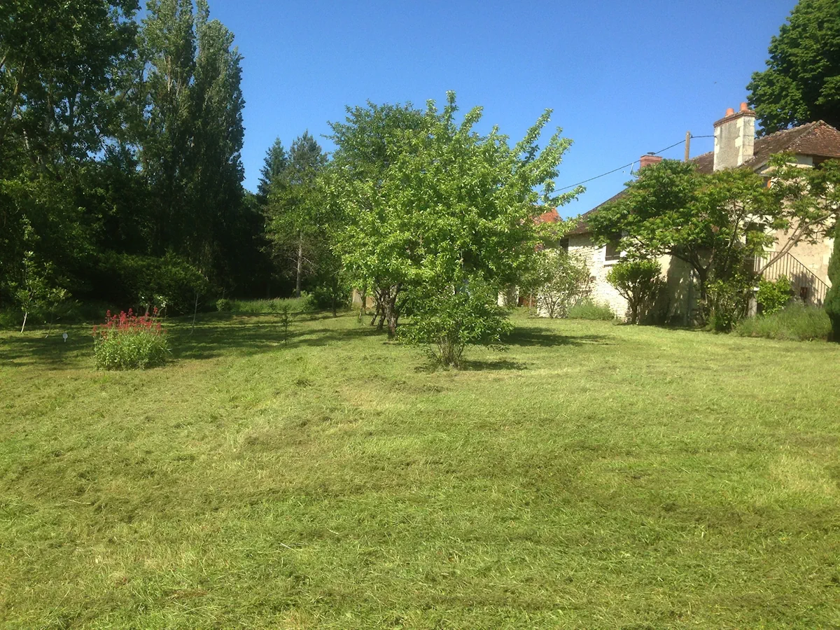 Verger fauché autour d'un pommier, maison de pierre et massifs en arrière-plan