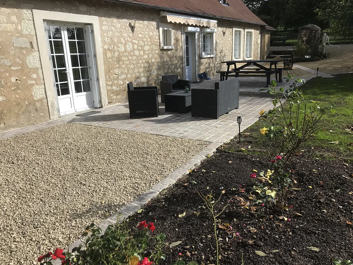 Pelouse et massif de rosiers bordant une terrasse pavée devant une longère de pierre
