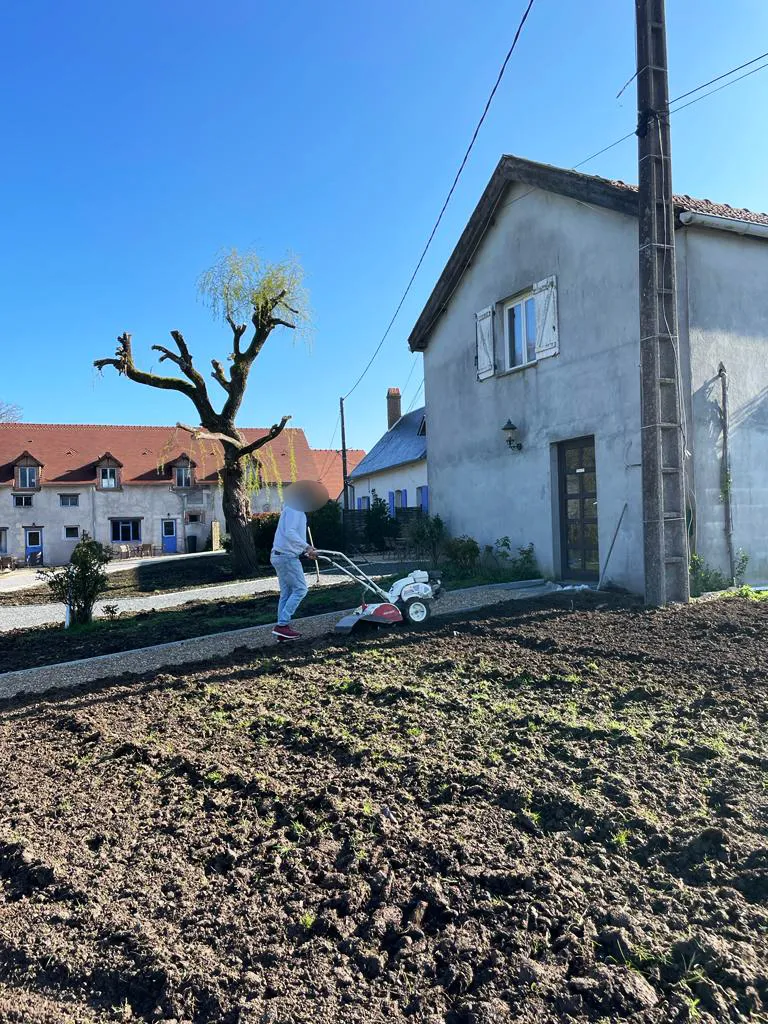 Compagnon passant le motoculteur sur une large bande de terre avant semis, devant une maison de village