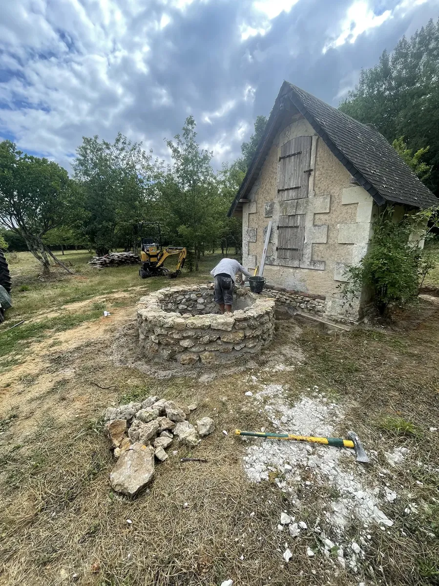 Margelle de puits en cours de montage dans un verger, mini-pelle et tas de pierre autour