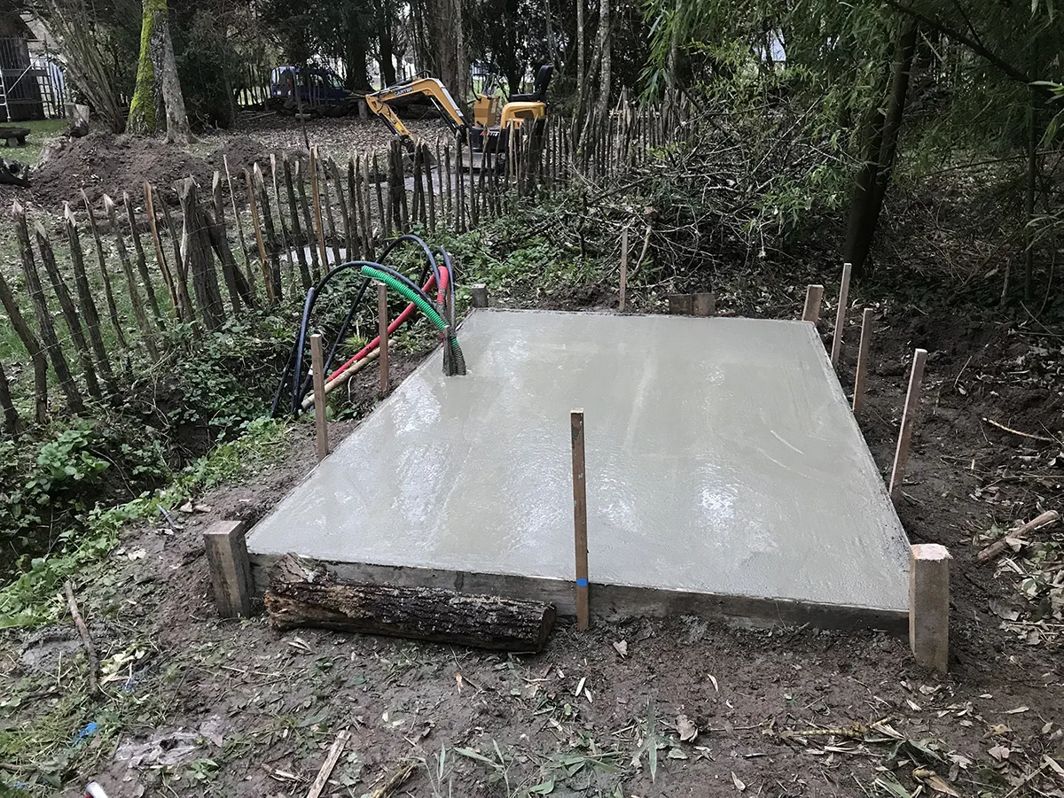 Dalle de béton fraîchement coulée et talochée entre des ganivelles, réseaux en attente