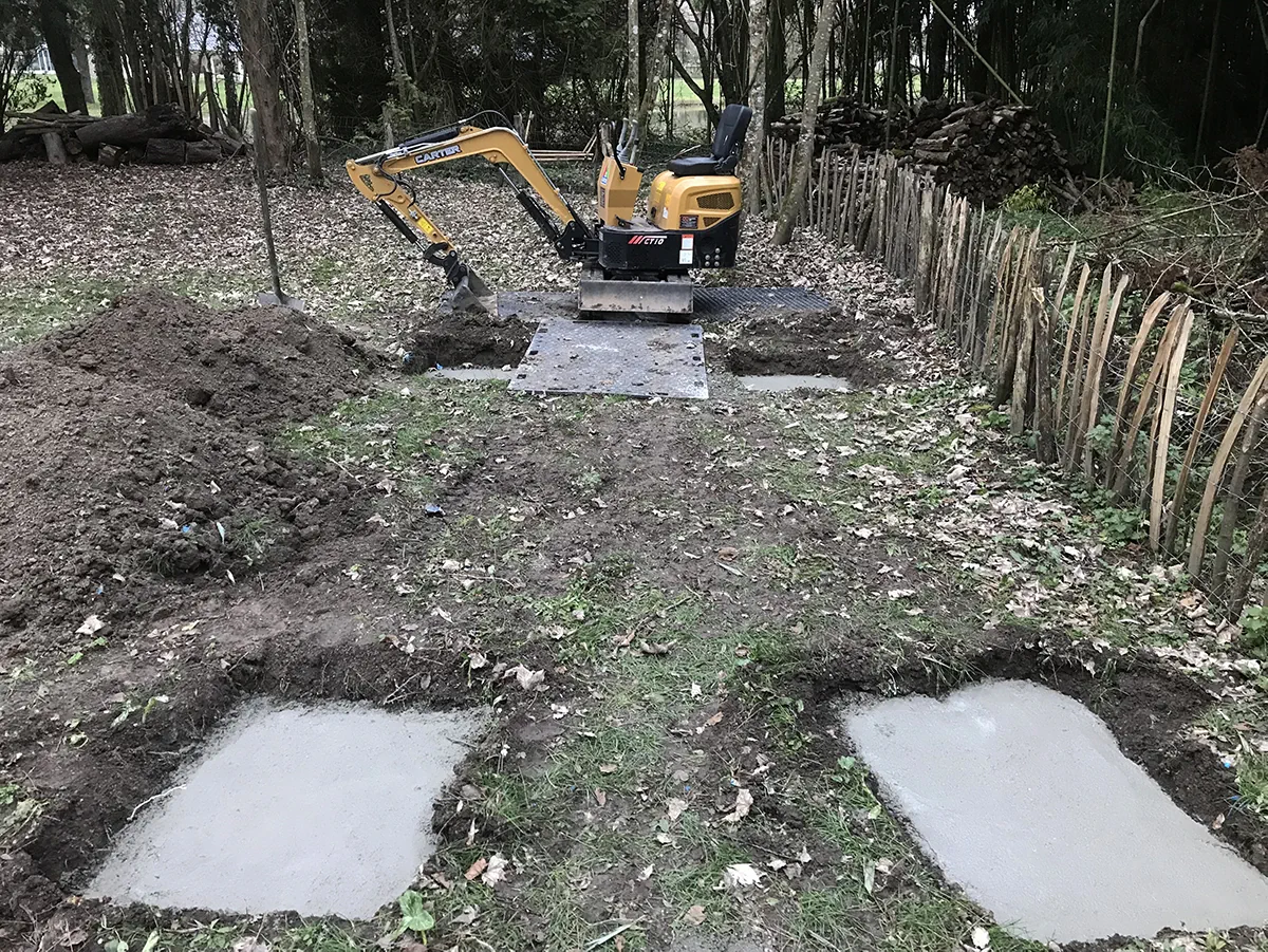 Plots de béton coulés en sous-bois pour recevoir une construction, mini-pelle posée sur ses plaques