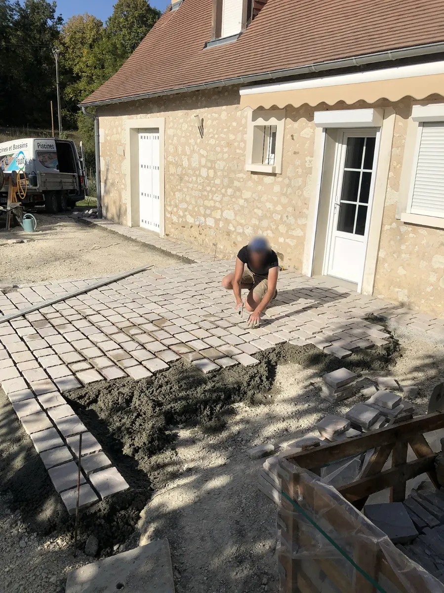 Compagnon posant à la main des pavés de pierre sur lit de mortier, devant une maison en pierre