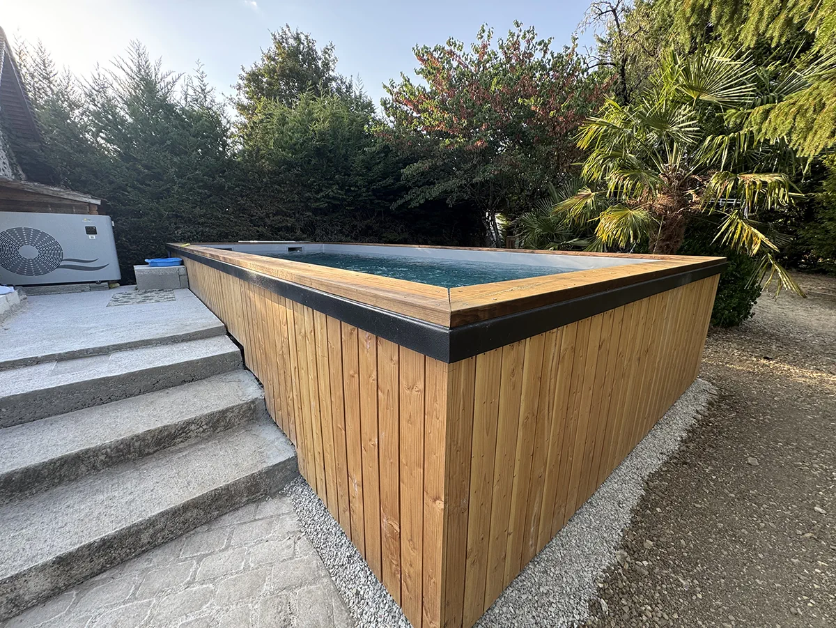 Piscine hors-sol habillée d'un bardage en lames de bois clair et coiffée d'une margelle noire, au pied d'un escalier de pierre