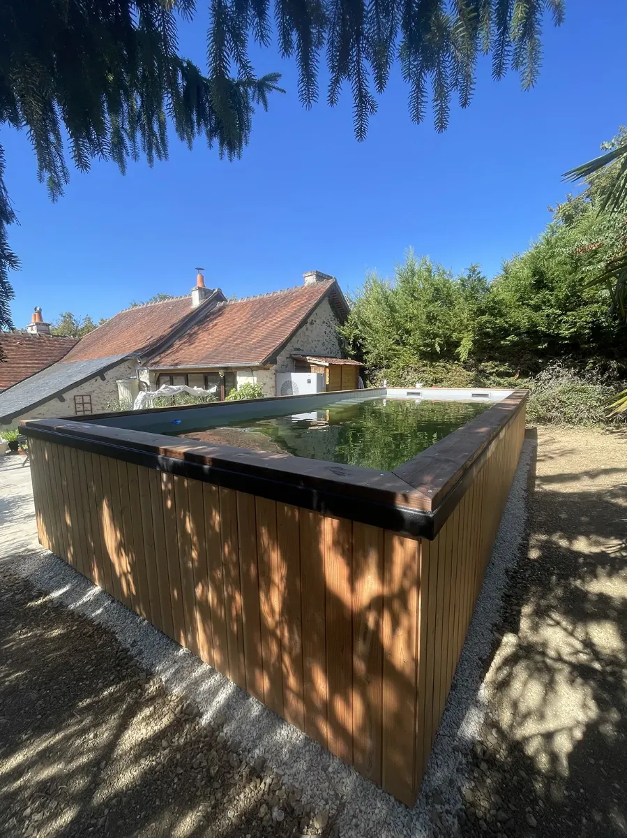 Piscine hors-sol au bardage de bois clair et à la margelle noire, devant une longère aux toits de tuile