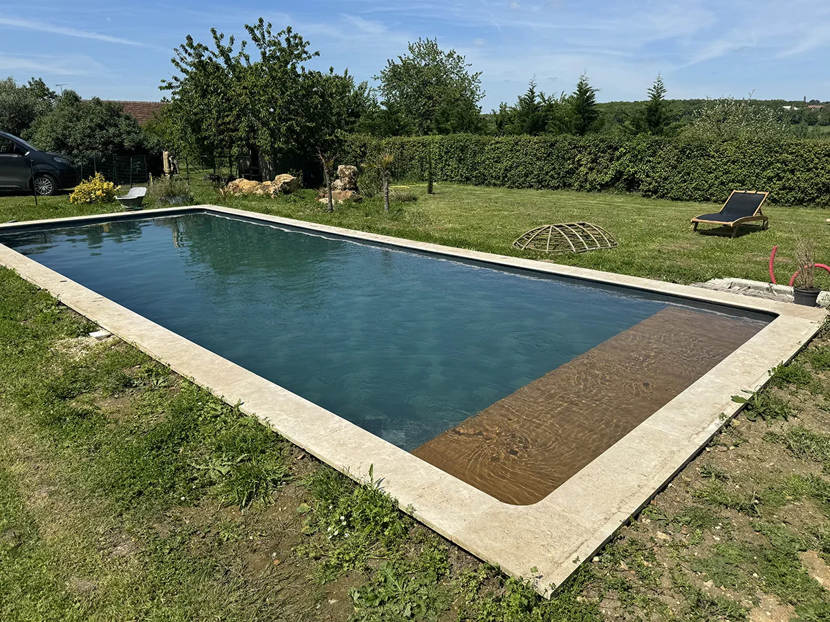 Grande piscine maçonnée à margelle de travertin, dans une prairie bordée d'une haie, sous un ciel d'été