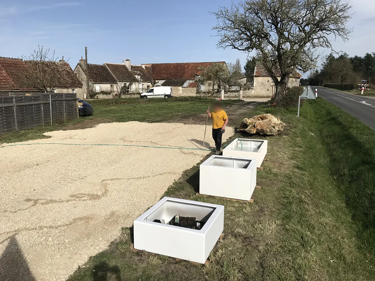 Trois bacs de béton blanc posés sur cales le long d'une aire de gravier, avant plantation