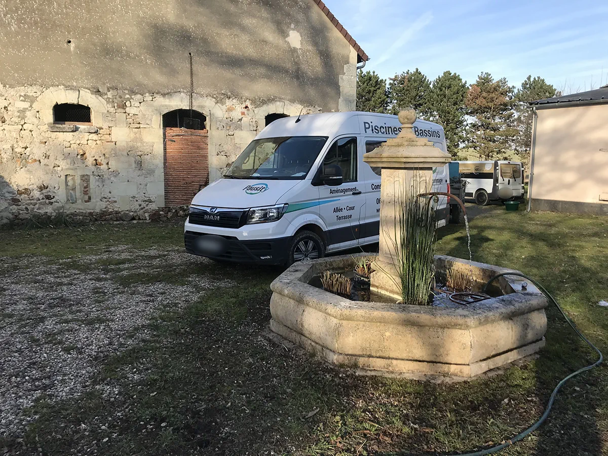 Ancienne fontaine de pierre remise en eau et plantée de joncs, fourgon Pausity garé devant une grange