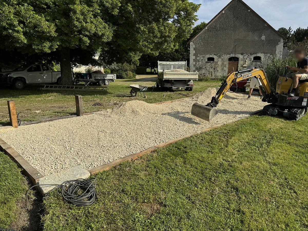 Mini-pelle étalant le gravier d'un terrain de boules en cour de ferme, camion-benne et hangar de pierre à l'arrière