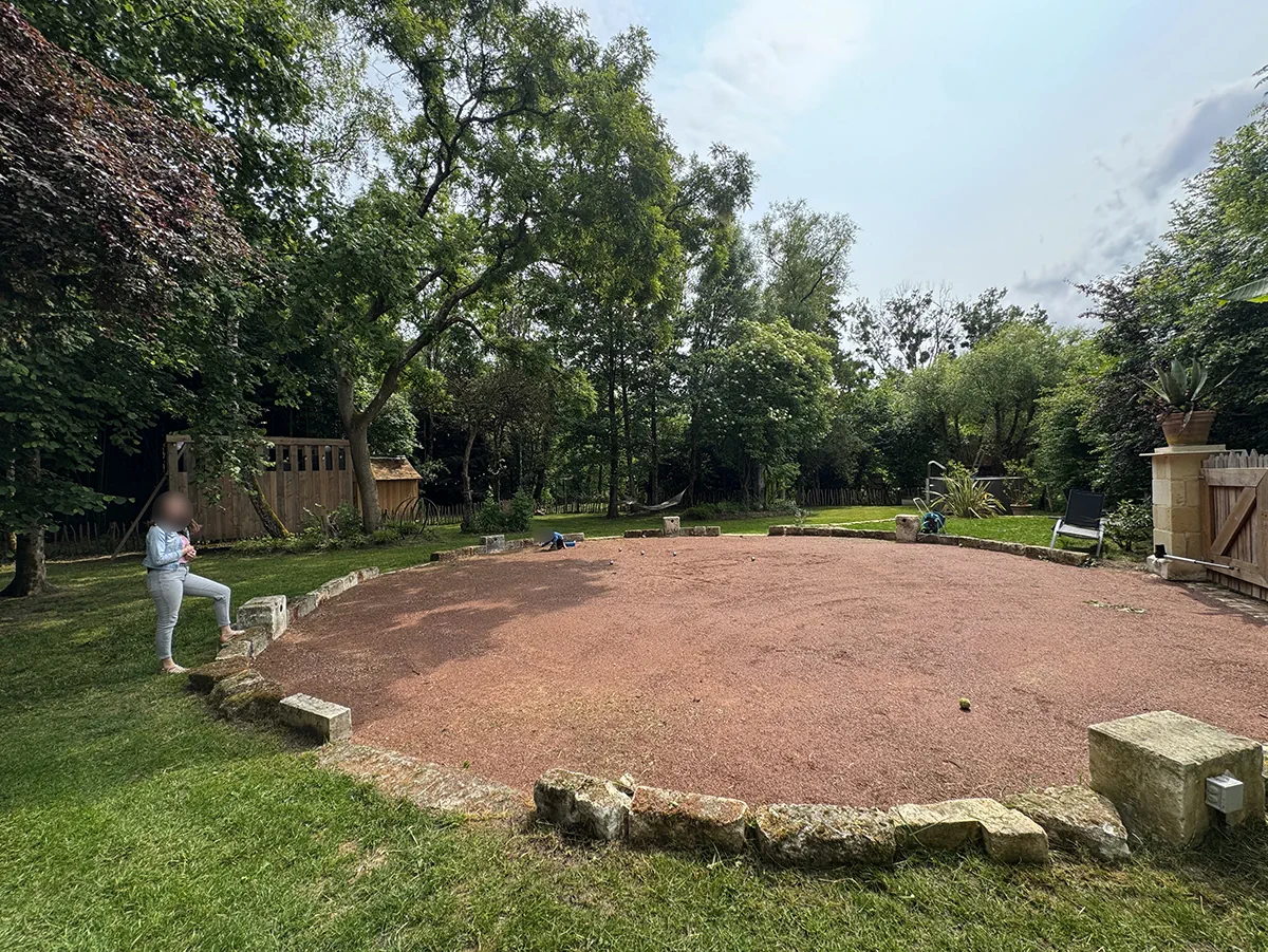 Terrain de boules de forme ovale en mâchefer rose, ceinturé de blocs de pierre, au milieu d'un jardin arboré