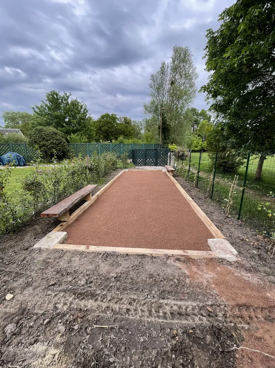 Terrain de boules vu dans sa longueur, bordures de madriers et blocs de pierre d'angle, banc de bois sur le côté