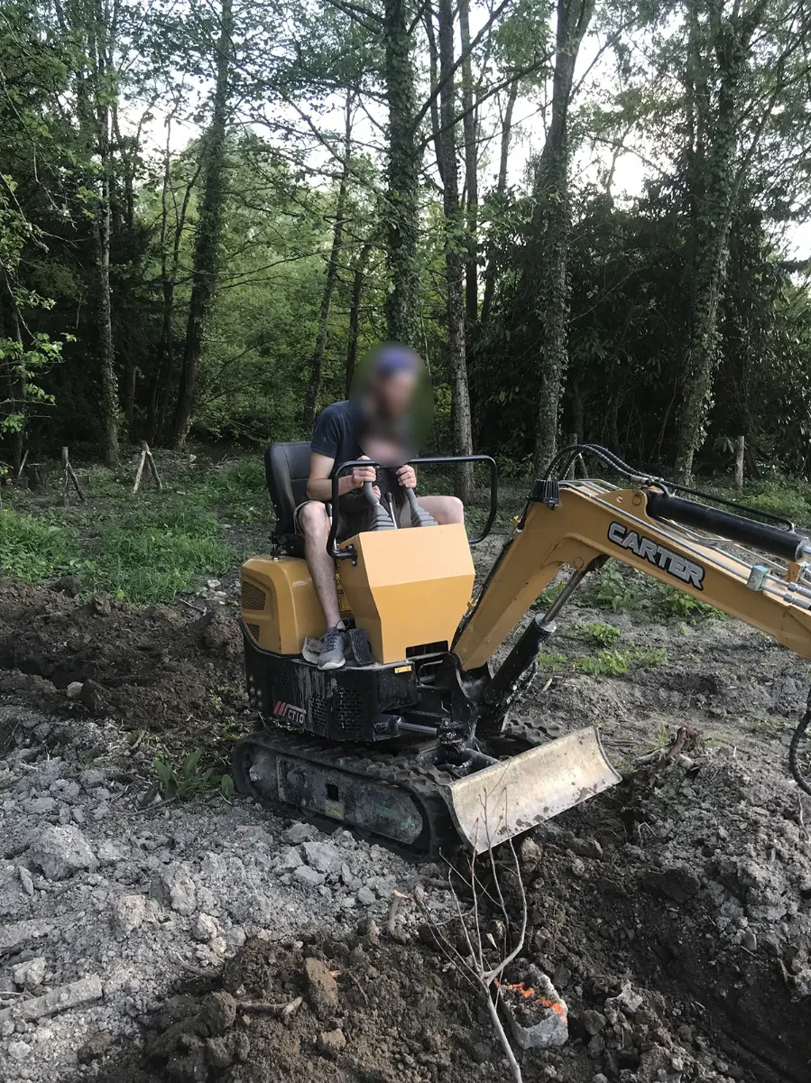 Compagnon aux commandes d'une mini-pelle sur un terrain décapé, en lisière de sous-bois