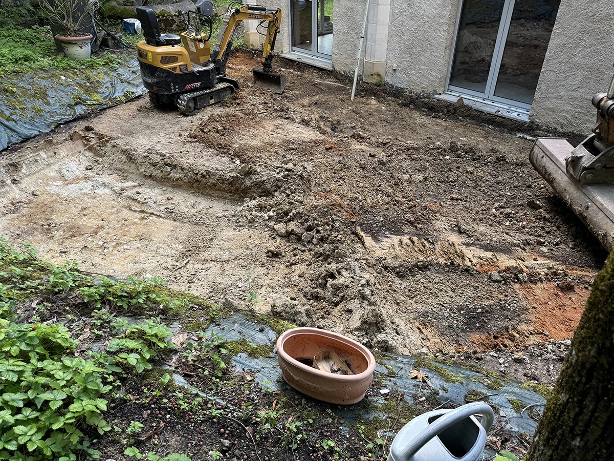 Mini-pelle décaissant une cour au pied d'une maison, terre et gravats mis à nu avant reprise