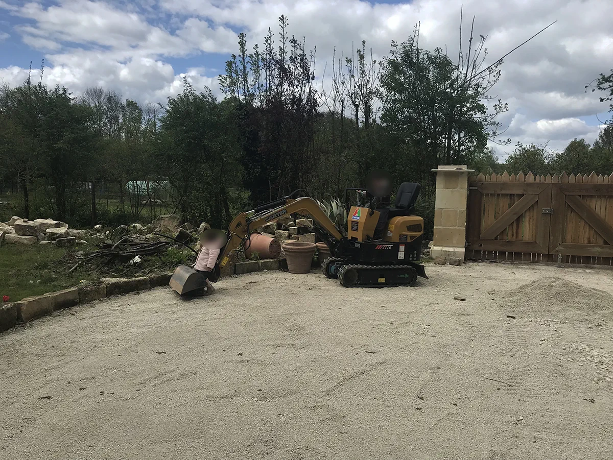 Mini-pelle à l'arrêt sur une cour de gravier fraîchement nivelée, devant un portail de bois