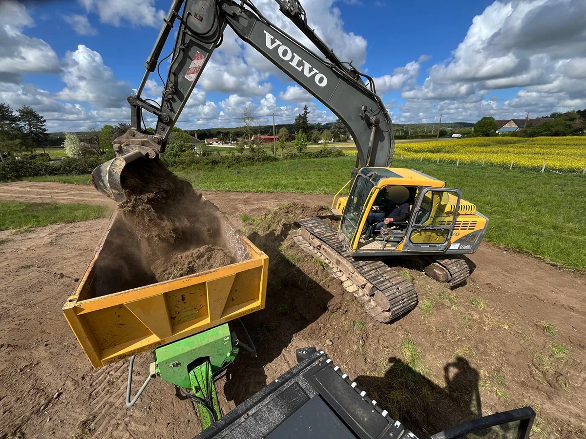 Pelle Volvo déversant un godet de terre dans une benne agricole, champ de colza en arrière-plan
