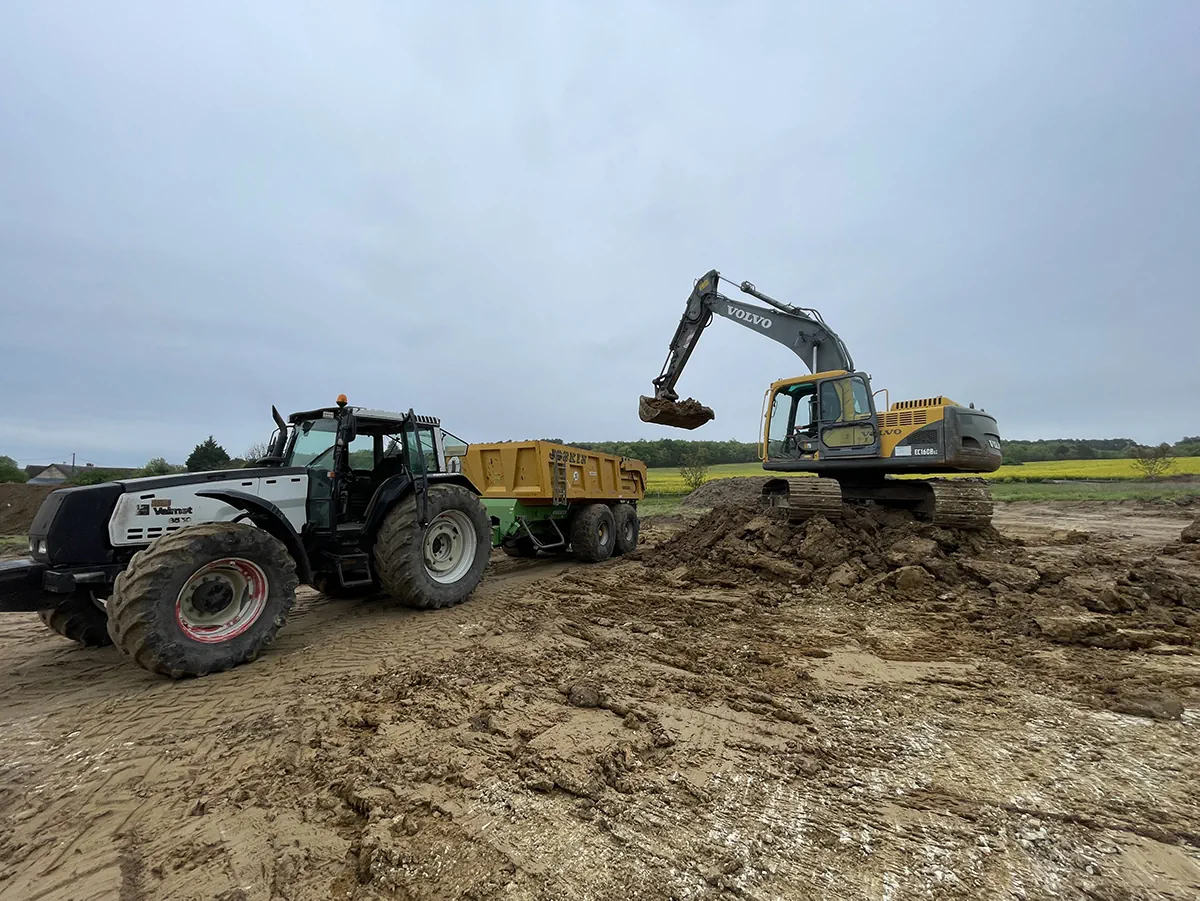 Tracteur attelé à une benne et pelle Volvo au travail sur un terrain détrempé, sous un ciel couvert