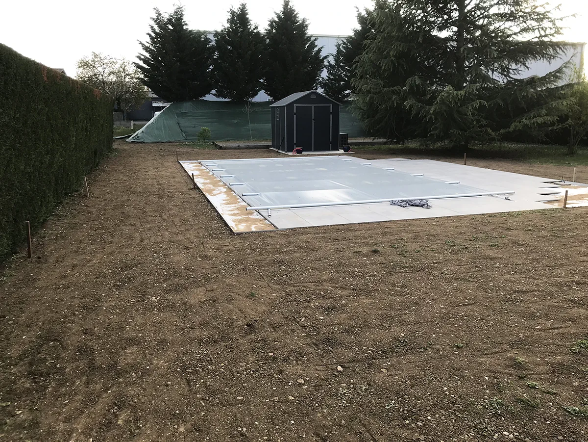 Dalle de béton coulée et bâchée sur un terrain nu, abri de jardin et haie de thuyas en fond