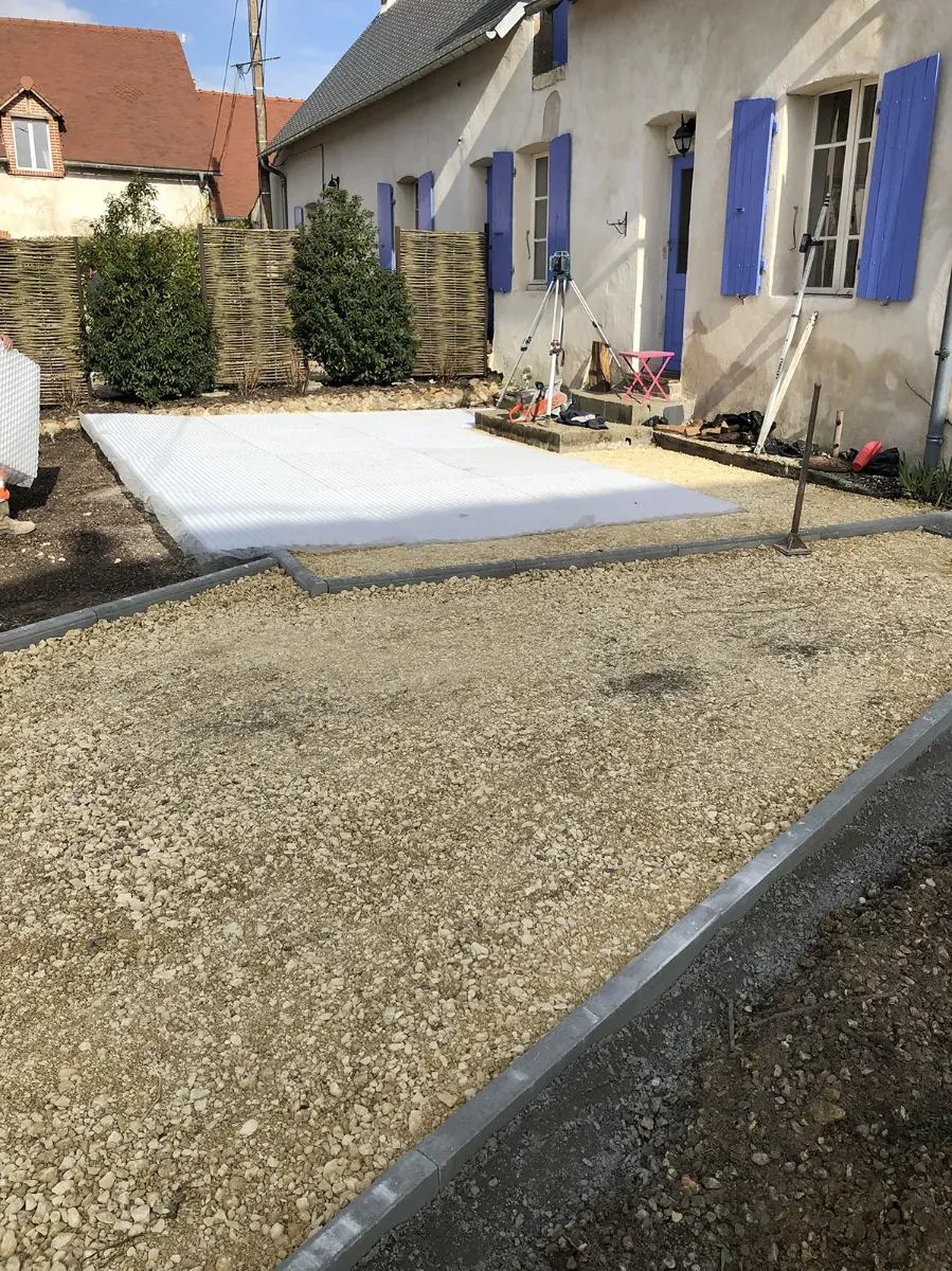 Géotextile déroulé et lit de pose préparé pour une terrasse, devant une maison aux volets bleus