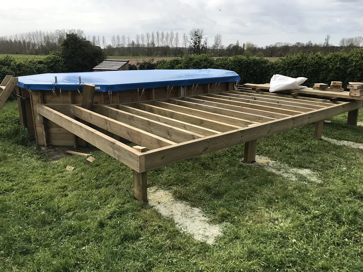 Ossature de terrasse en bois montée contre une piscine hors-sol bâchée, en bordure de champ