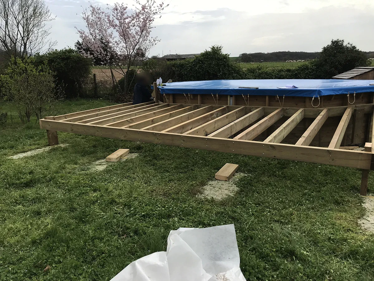 Ossature de terrasse en bois montée sur poteaux le long d'une piscine bâchée, dans une prairie