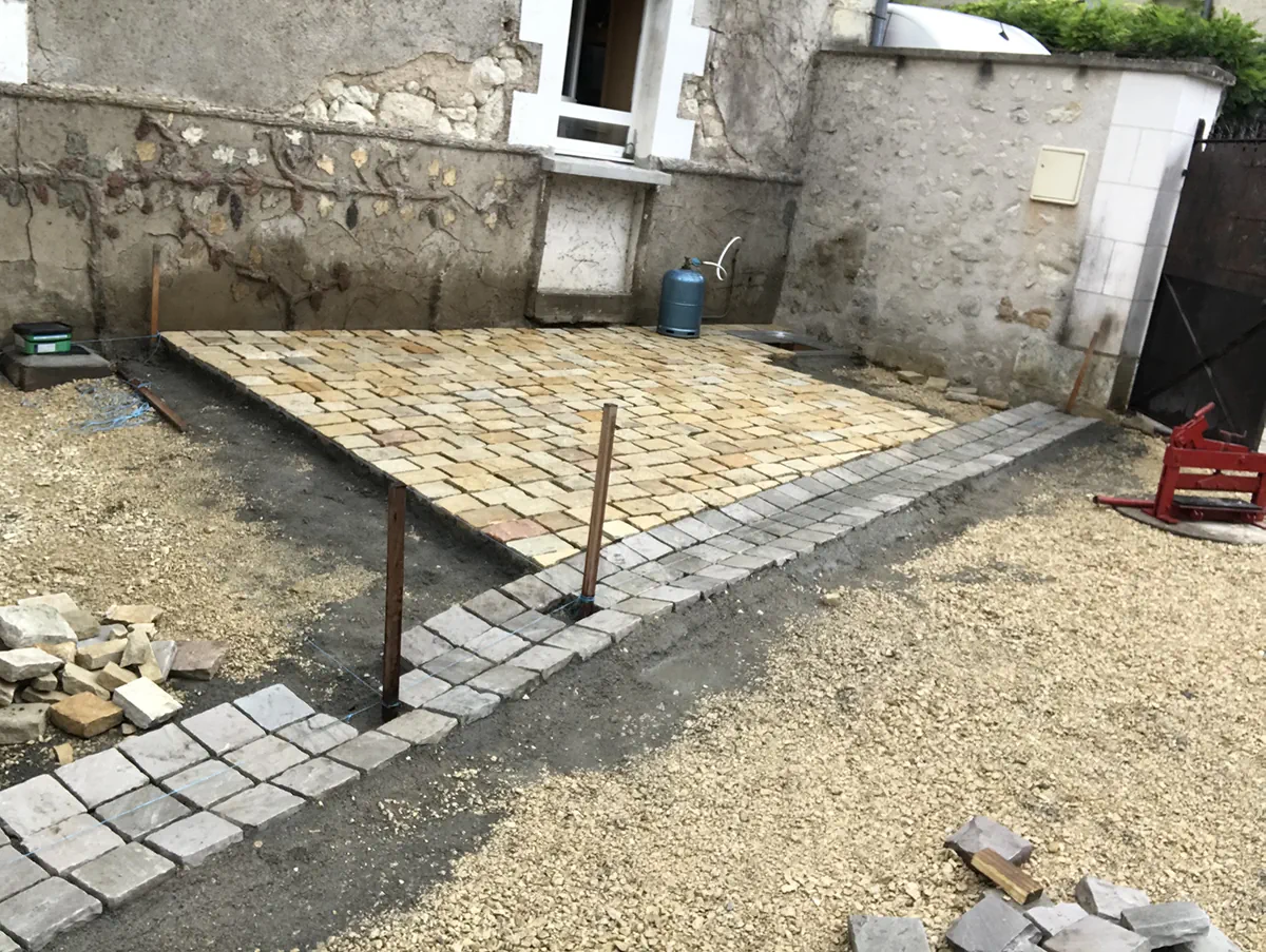 Terrasse en pavés de pierre dorée en cours de pose dans une cour, bordure grise et piquets de niveau