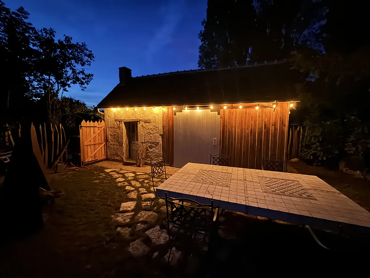 Terrasse carrelée devant une grange de pierre, éclairée par une guirlande à la tombée de la nuit
