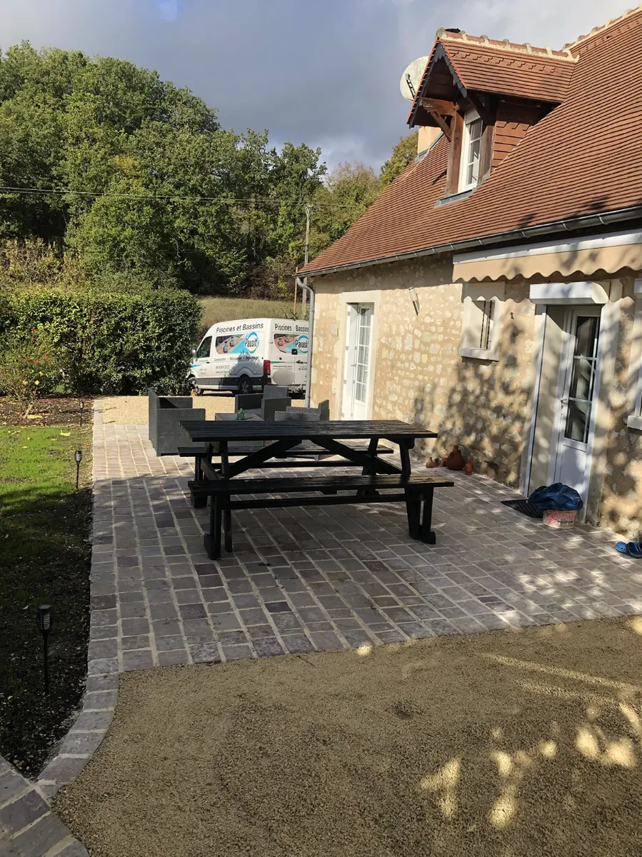 Terrasse en pavés de pierre et table de jardin devant une longère, fourgon Pausity garé à l'arrière