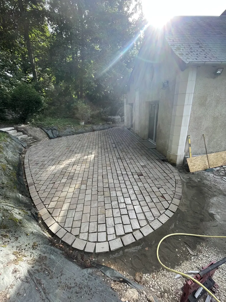 Grande terrasse en pavés de pierre au tracé courbe, posée devant une maison, dans la lumière rasante du matin